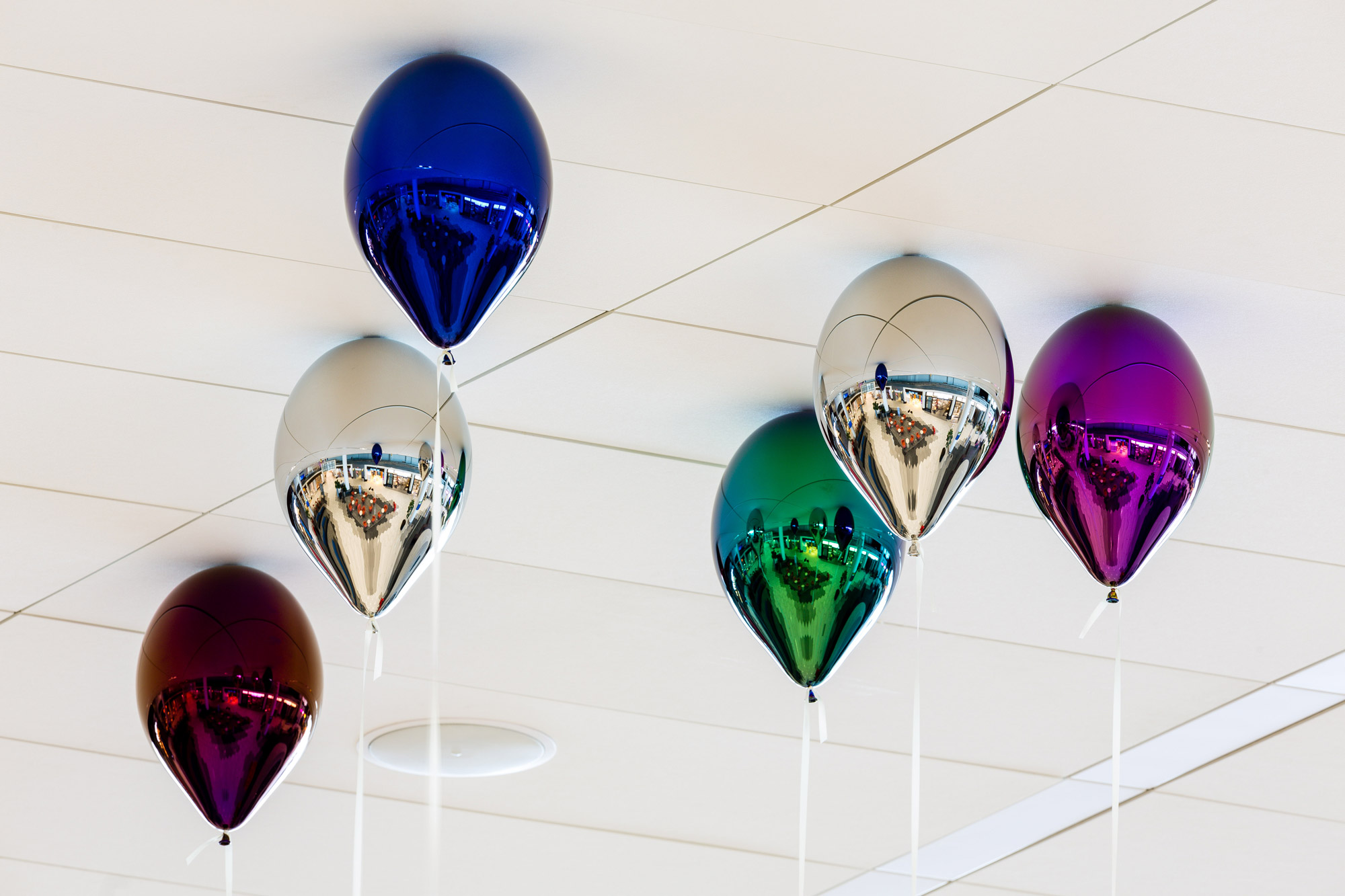 A series of metallic balloons appear to float against a white ceiling. The balloons are in blue, silver, green, magenta, red, and silver.
