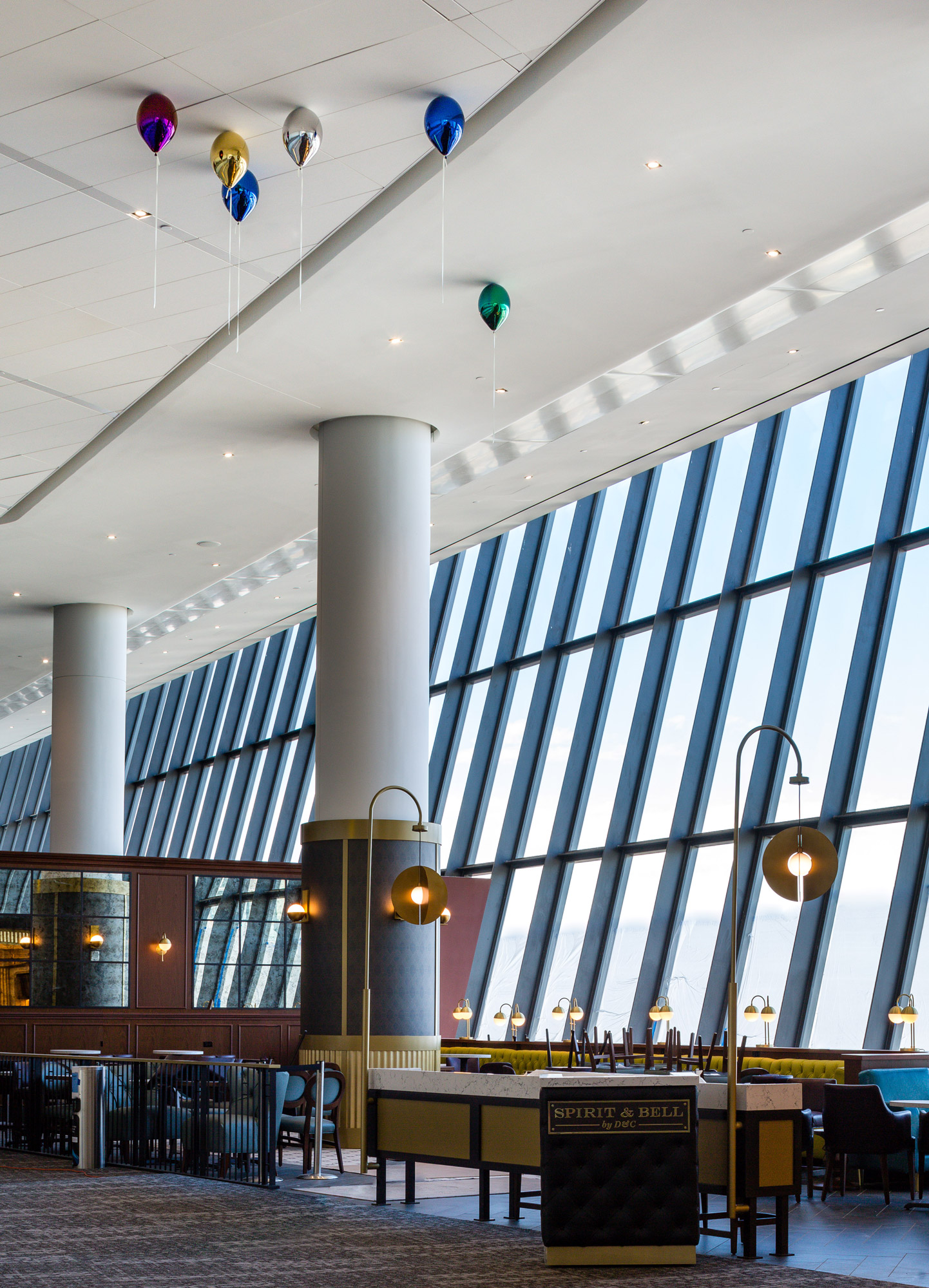 People walk through an airport terminal, underneath tall white ceilings where a series of colorful metallic balloons appear to float.