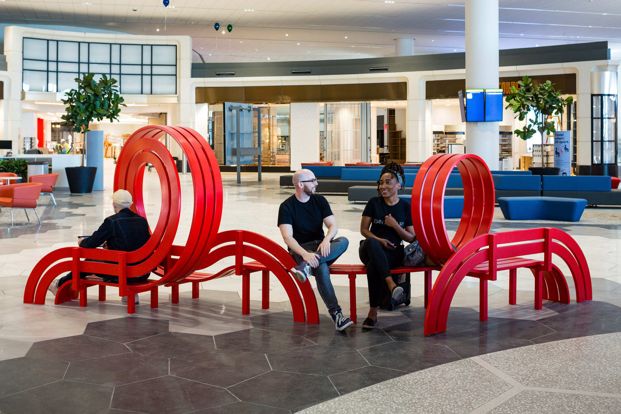 A red sculptural bench and set of metallic colorful balloons pictured in an airport terminal with people milling about.