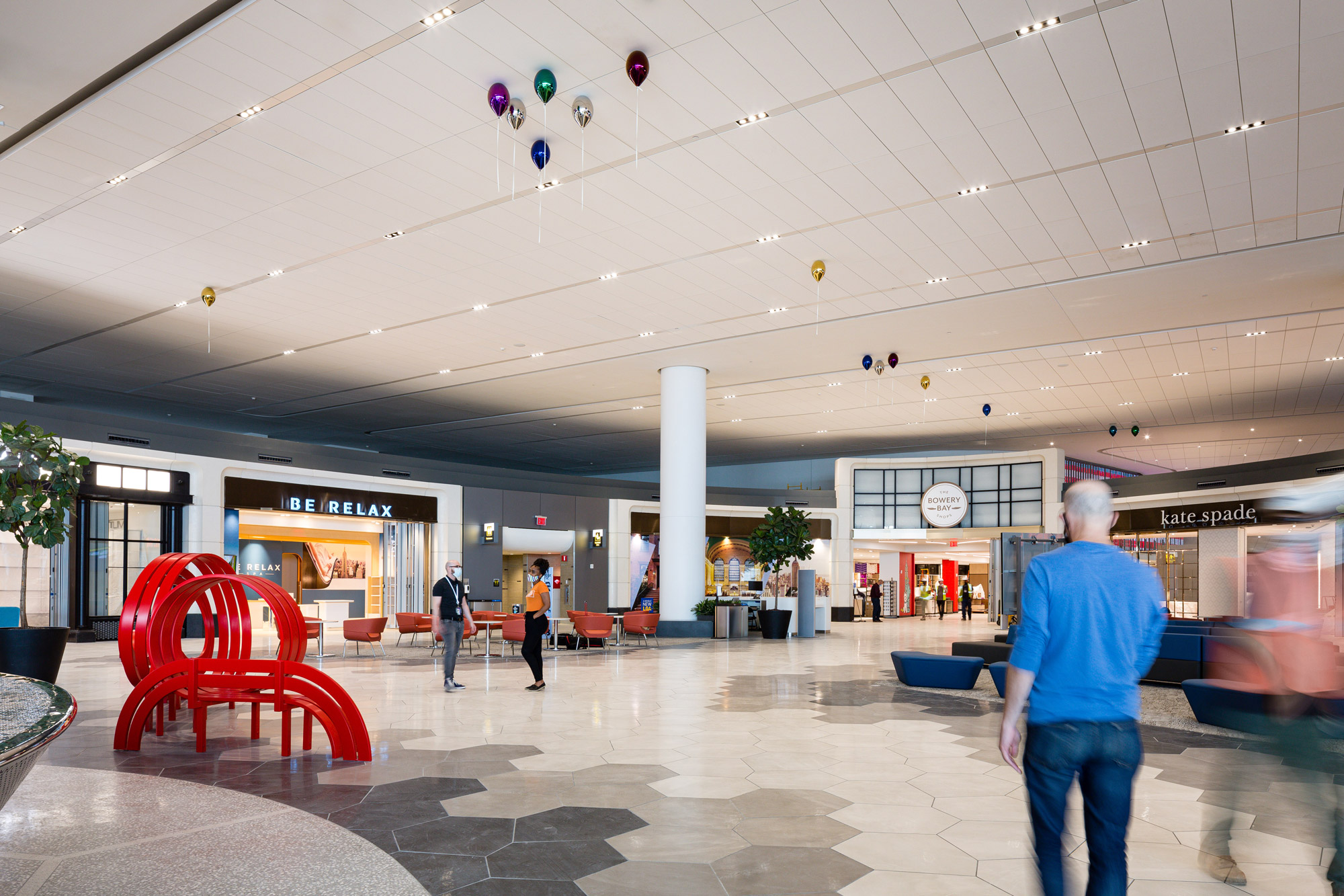 A red sculptural bench and set of metallic colorful balloons pictured in an airport terminal with people milling about.