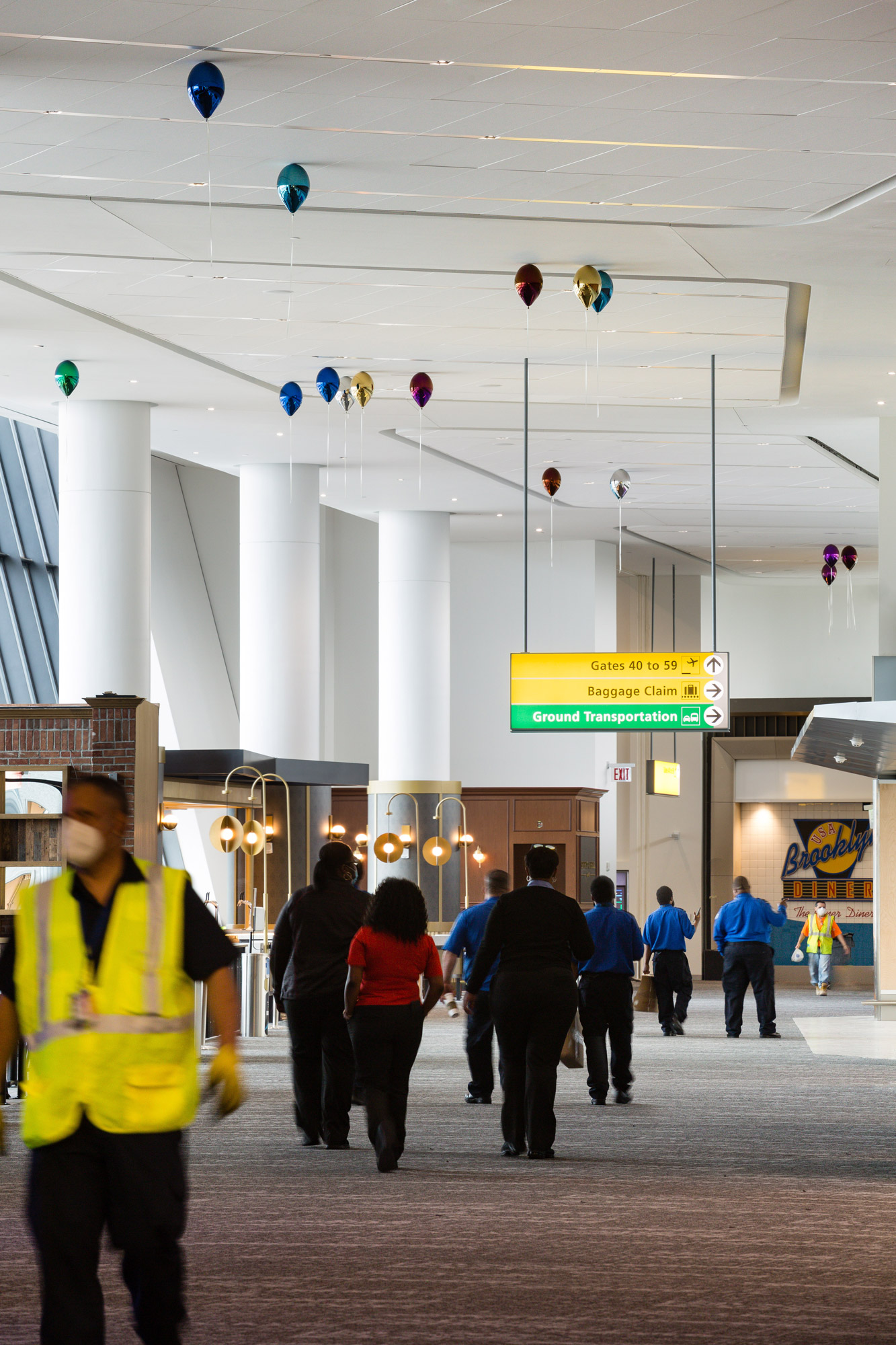 People walk through an airport terminal, underneath tall white ceilings where a series of colorful metallic balloons appear to float.