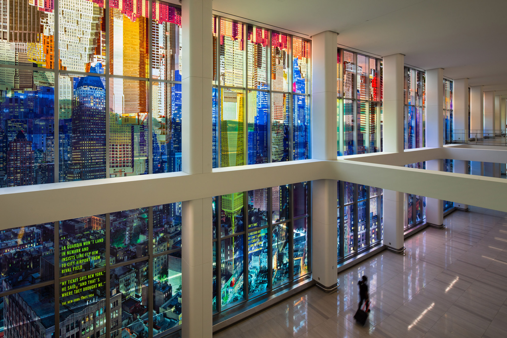 A kaleidoscopic wash of a colorful photo-collaged cityscape fills the length of two stories of tall glass windows in an airport terminal interior.
