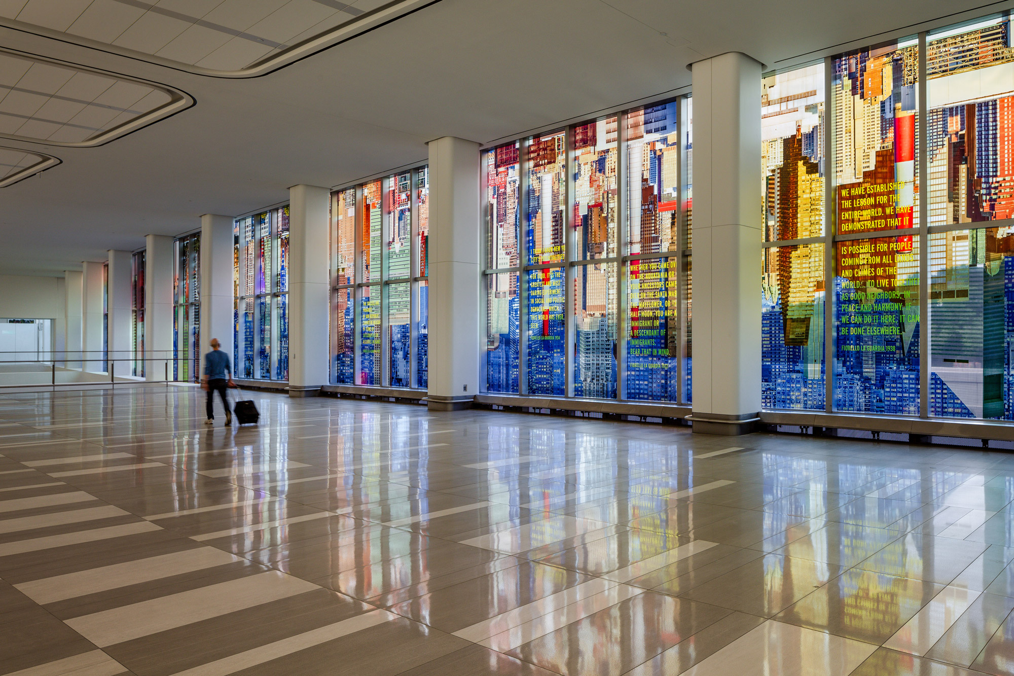 A kaleidoscopic wash of a colorful photo-collaged cityscape fills the length of two stories of tall glass windows in an airport terminal interior.