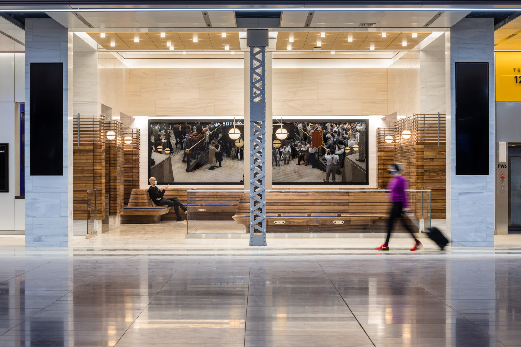 A wall of a train lobby houses two horizontal panels of photographs, with blue upholstery and wood seating below the panels. The two photographs depict reenactments...