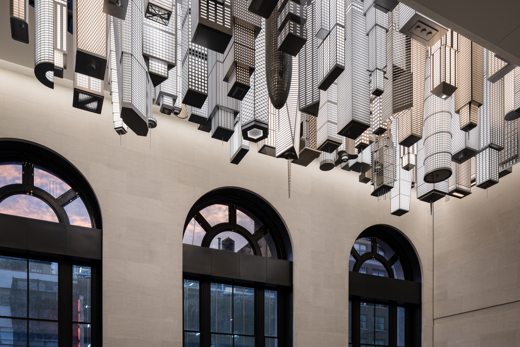 A crop of black and white lit up high-rise buildings descending from the ceiling of the inside of a train station, with three arched windows.