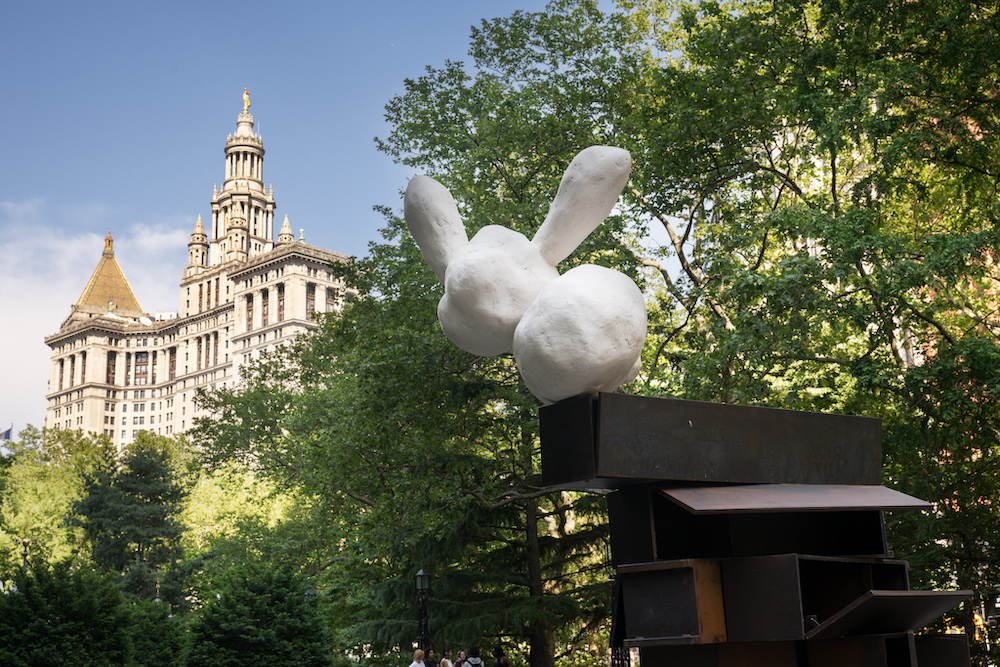 A round rabbit-ear resembling white form balances atop a precarious looking steel structure, with a city building and foliage in the background.