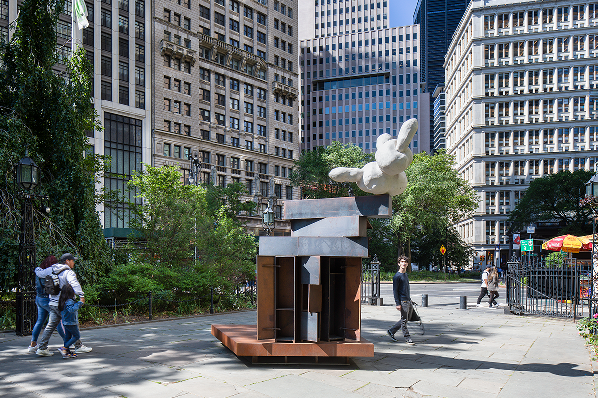 A round rabbit-resembling white form balances atop a steel structure resembling stacked shelves, with high-rise buildings in the background.