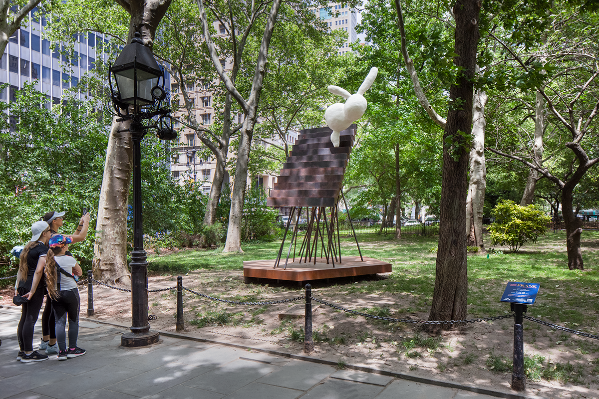 A round rabbit-resembling white form balances atop a steel stair structure in a park surrounded by trees. Three onlookers snap a photo from the walkway.