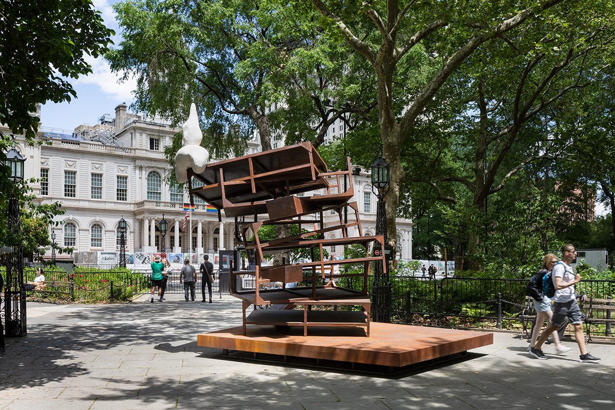 A round rabbit-resembling white form balances atop a steel structure resembling stacked tables in a park, set against a city hall building.