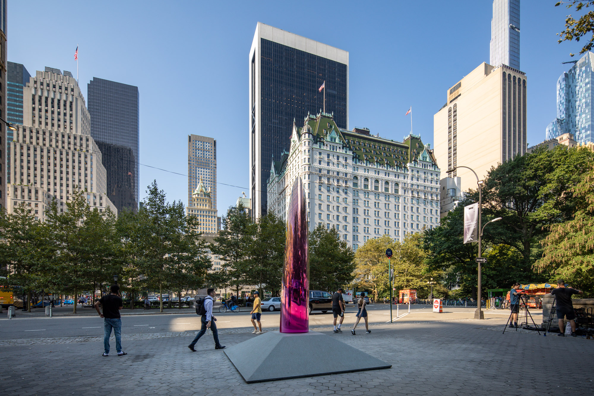 A 12-foot tall translucent and reflective magenta parabolic sculpture inhabits a park in a bustling city.