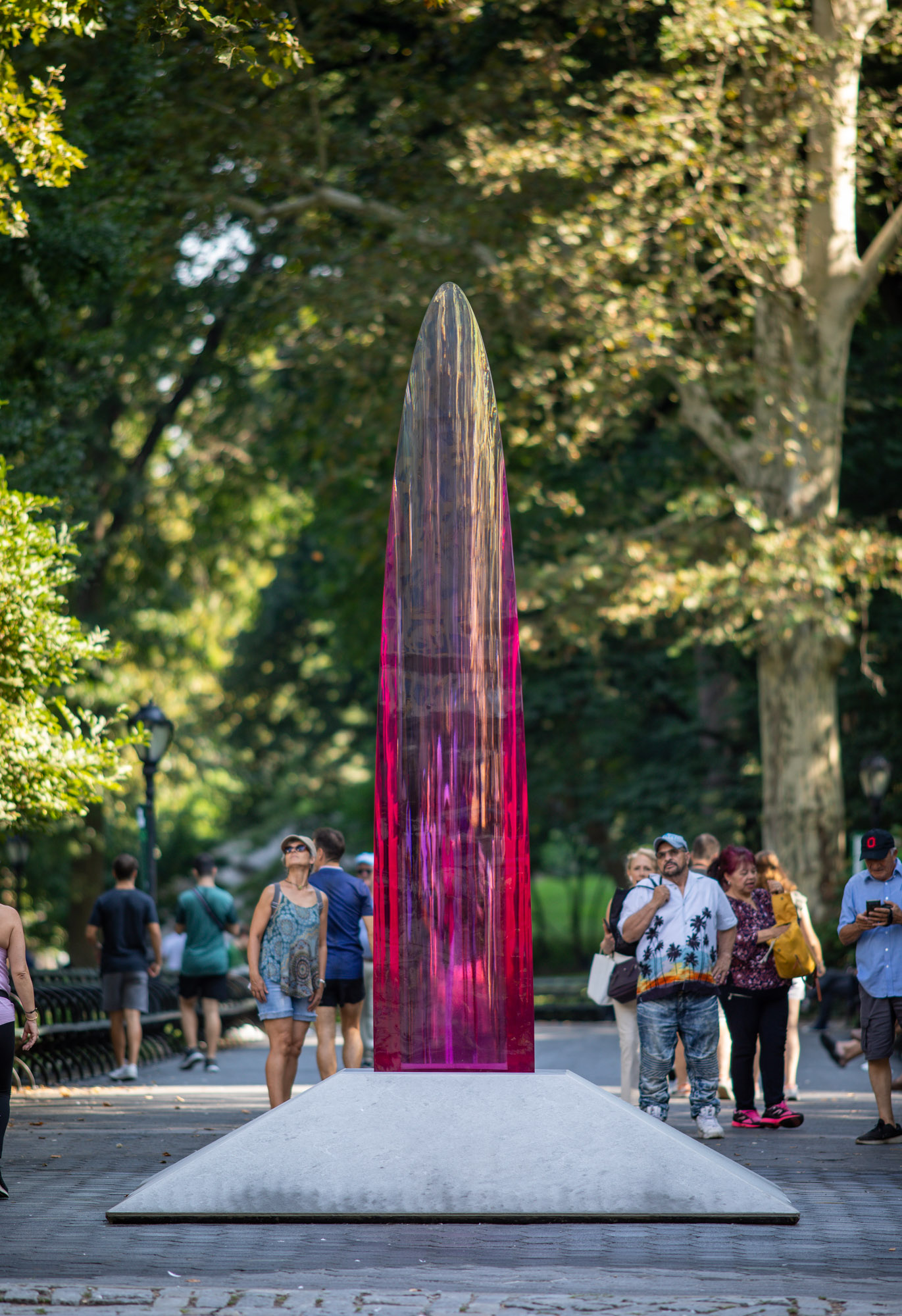 A 12-foot tall translucent and reflective magenta parabolic sculpture inhabits a park in a bustling city. several visitors gaze up at the work.