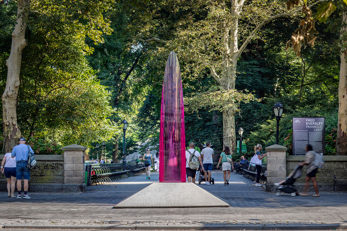 A 12-foot tall translucent and reflective magenta parabolic sculpture inhabits a park in a bustling city.