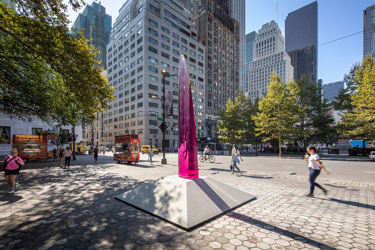 A 12-foot tall translucent and reflective magenta parabolic sculpture inhabits a park in a bustling city.