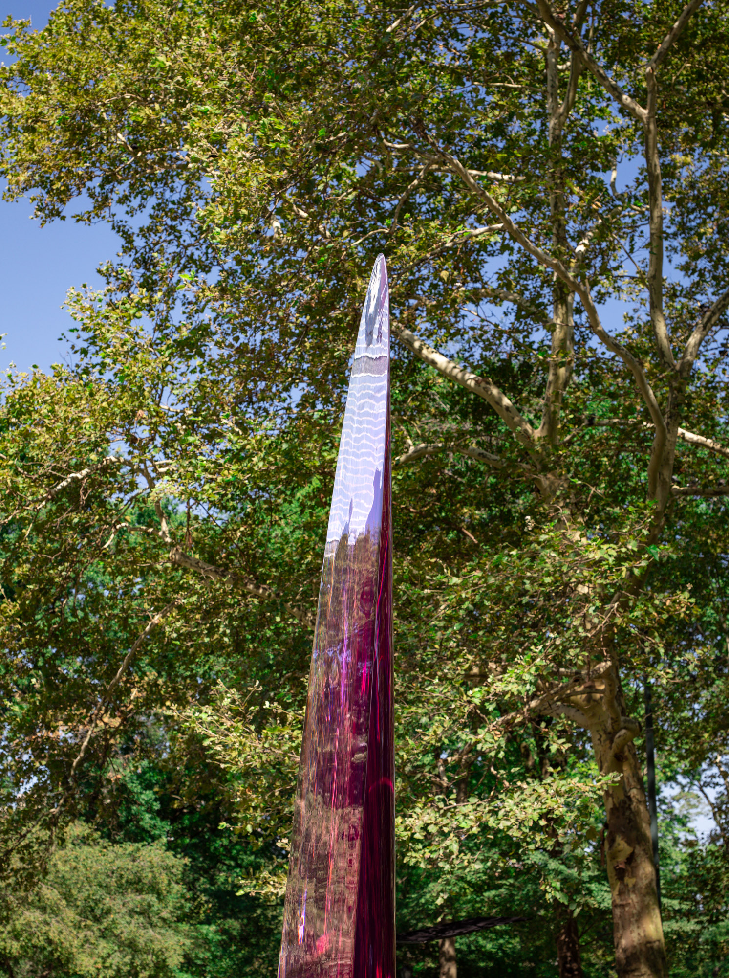 A 12-foot tall translucent and reflective magenta parabolic sculpture inhabits a park in a bustling city.
