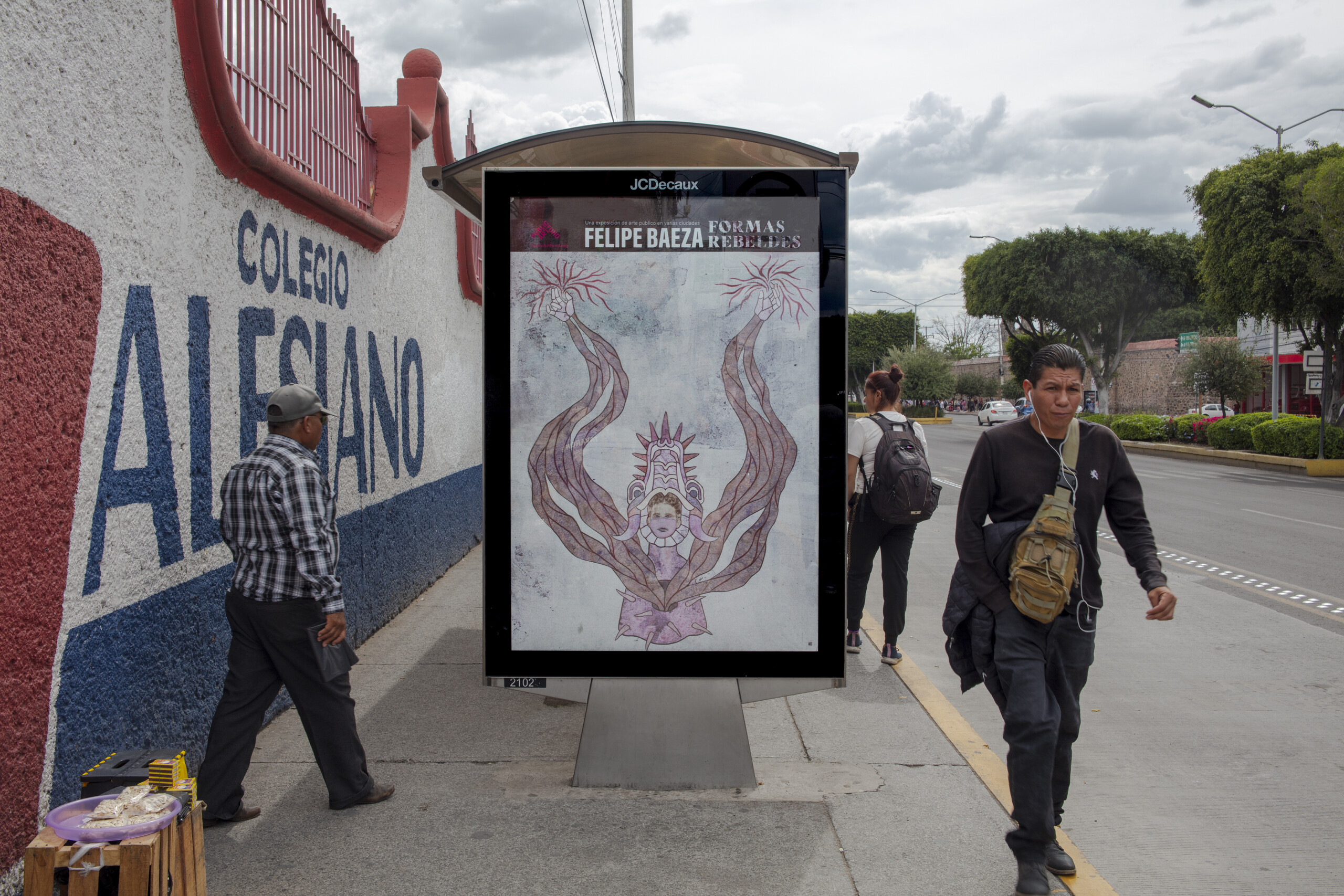 Pictured on a bus shelter on a busy city street is a layered hybrid figure that merges human, artifact, animal, and vegetation.