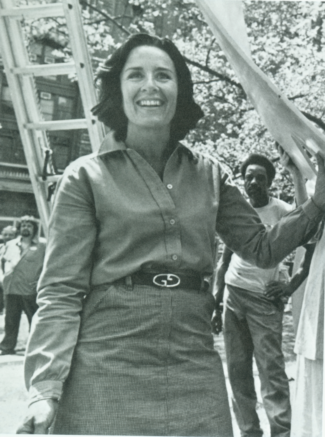 A black and white photo of a woman standing in a park, smiling.
