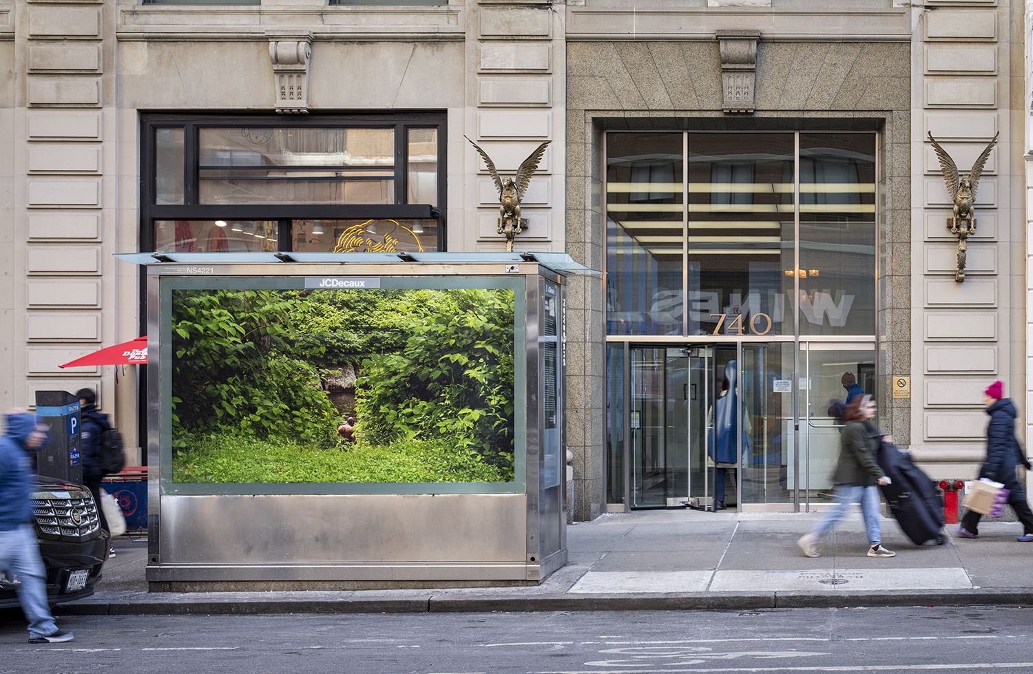 Pictured on a newsstand on a busy city street is a photograph of a Black man with his back facing the camera, barely visible amidst luscious green foliage. He appears...
