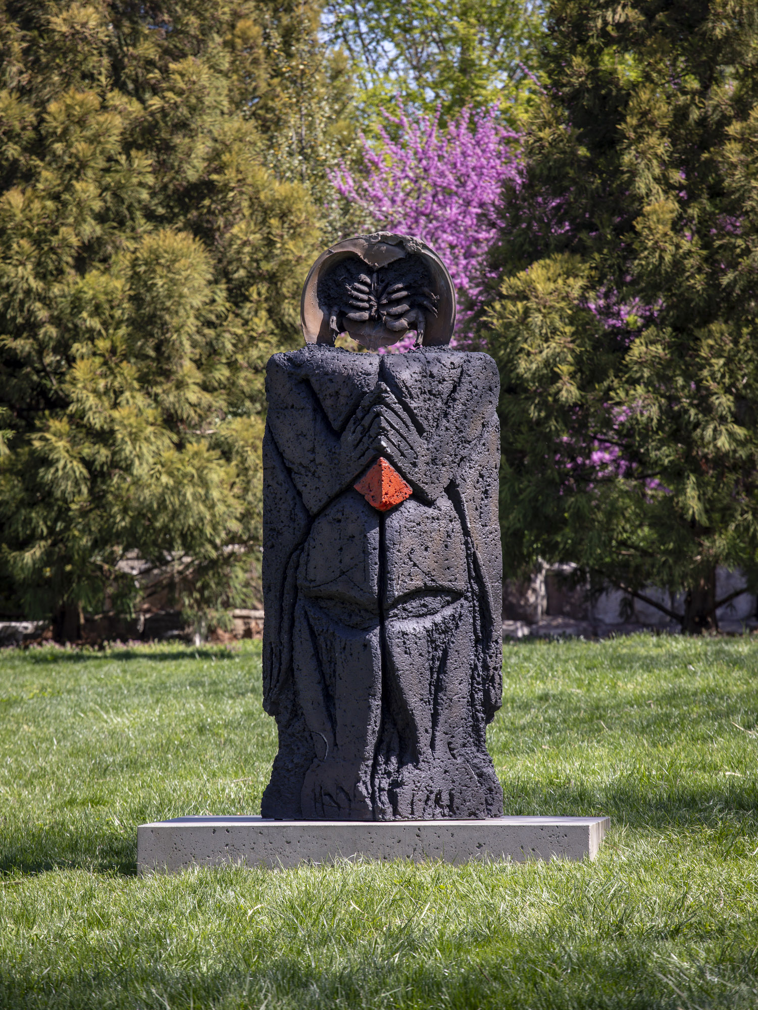 A close-up shot of a patinated bronze sculpture with a square concrete base installed along a grassy terrace. There is a spot of red towards the midpoint of the...