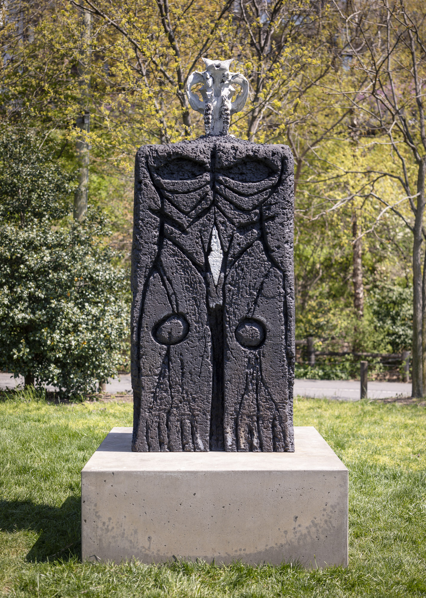 A close-up shot of a patinated bronze sculpture with a square concrete base installed along a grassy terrace. There are spots of yellow and white across the sculpture...