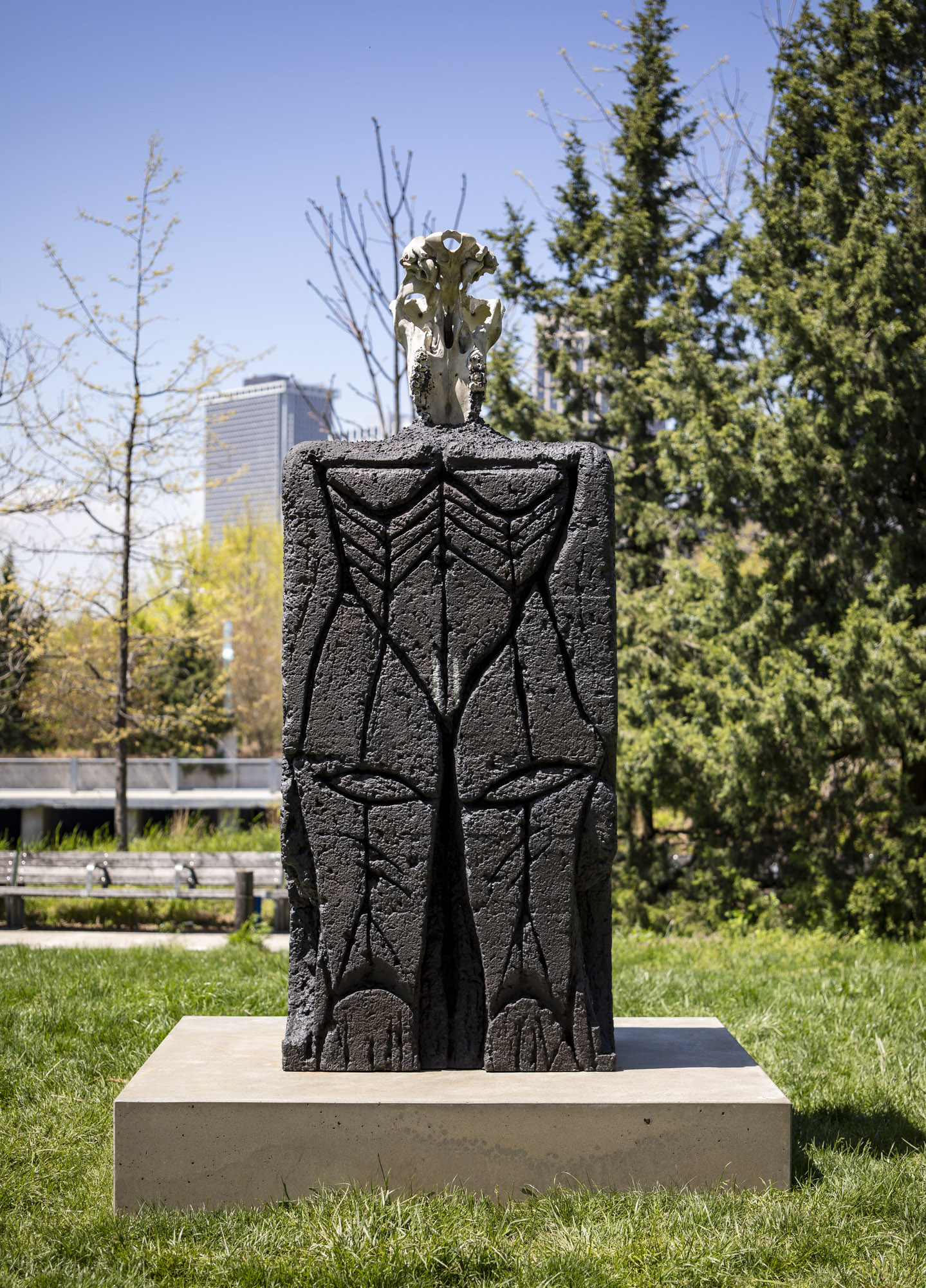 A close-up shot of a patinated bronze sculpture with a square concrete base installed along a grassy terrace. On top of the sculpture sits a recreated off-white...
