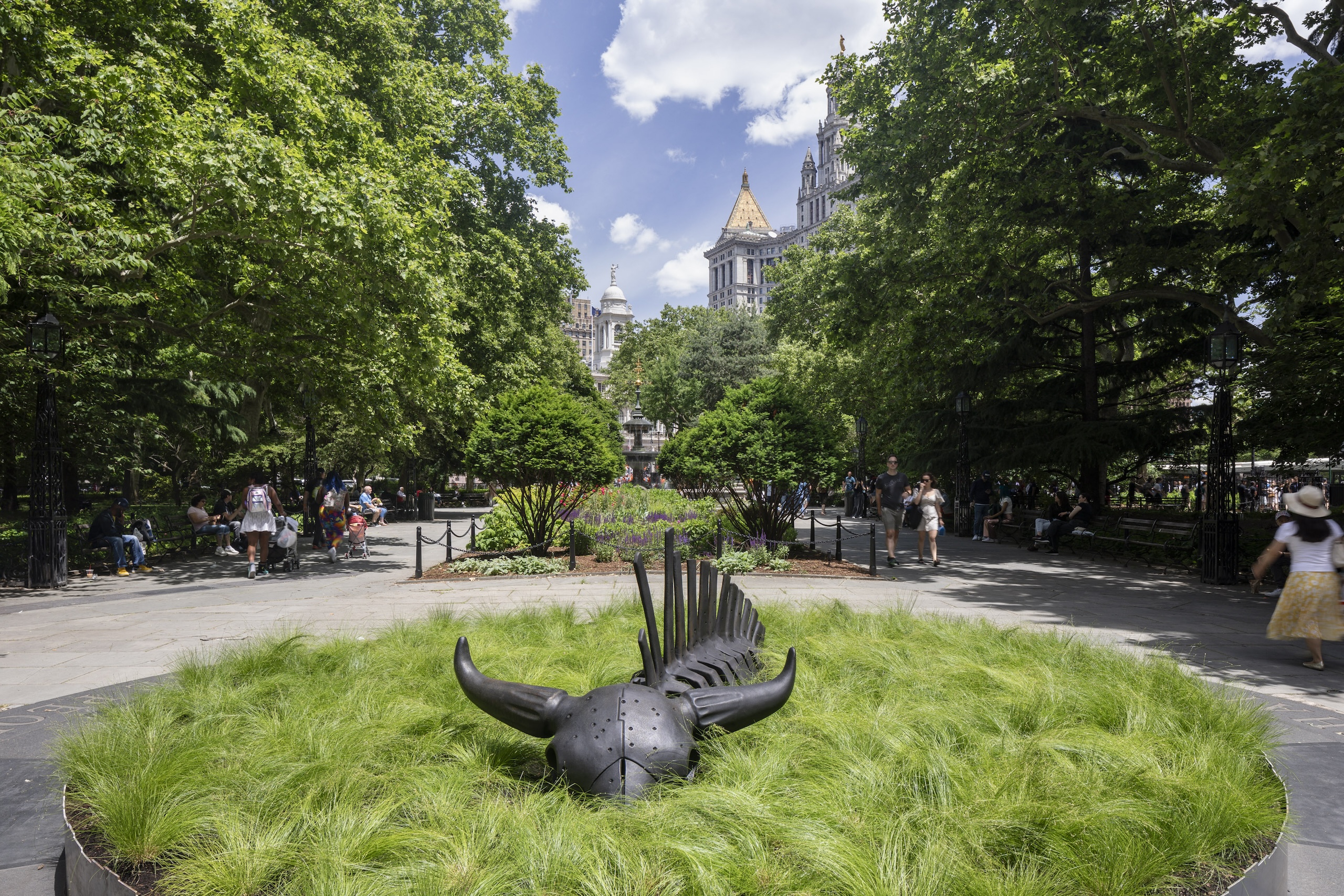 A 10-foot-long black steel sculpture of a bison skeleton sits within a shallow circular bed of green grass in a public park in New York City.